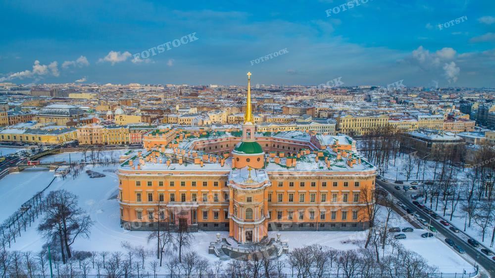 На фотографии запечатлен зимний день в Санкт-Петербурге. В центре изображения — здание Михайловского замка с его знаменитым золотой шпилем. Вокруг замка простираются парковые аллеи, покрытые снегом, и улицы, по которым движутся автомобили. Дальше видны крыши и фасады городских зданий, создающие колоритный городской пейзаж. 
Такой снимок может быть использован в культурно-исторических изданиях, для путеводителей и рекламы туристических маршрутов, а также как украшение в архитектурных и дизайнерских проектах.